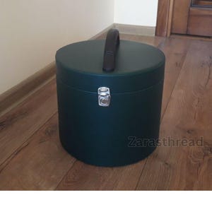 May include: A dark teal, cylindrical hat box with a black handle and a silver clasp. The box is on a wooden floor. The text "Zarasthread" is visible on the side of the box.