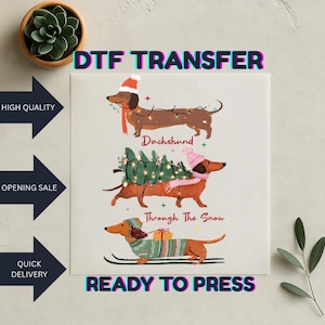 May include: A DTF transfer with three dachshund illustrations in Christmas outfits. The top dog has a Santa hat and lights, the middle dog carries a tree, and the bottom dog is on skis. Text includes "Dachshund" and "Through The Snow."