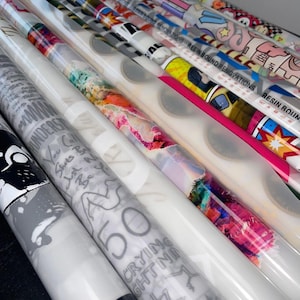 May include: A collection of rolled decorative papers with various designs. Some rolls feature colorful abstract art, while others display text and graphic illustrations. The rolls are wrapped in clear plastic, and some have the text "RESIN BOUND RENOVATIONS".