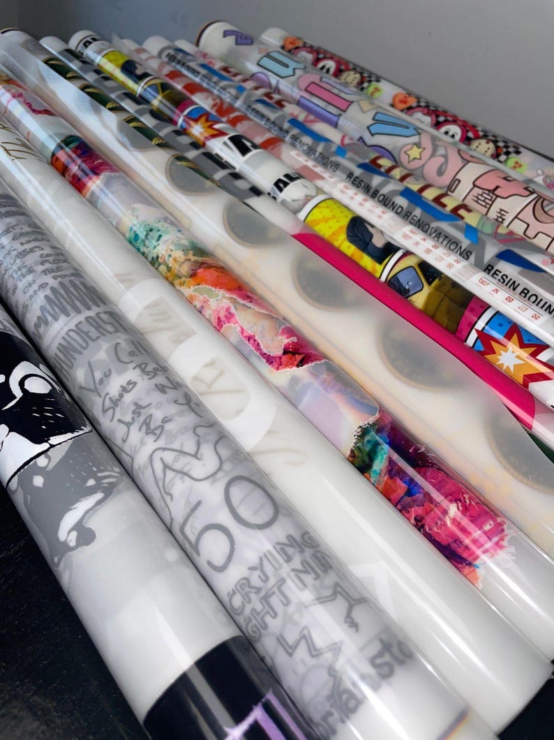 May include: A collection of rolled decorative papers with various designs. Some rolls feature colorful abstract art, while others display text and graphic illustrations. The rolls are wrapped in clear plastic, and some have the text "RESIN BOUND RENOVATIONS".