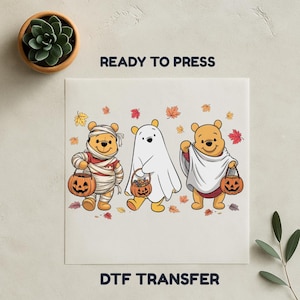 Op de afbeelding: Een Halloween DTF-transfer met drie stripberen in kostuums. Eén is in verbanden gewikkeld, een andere is een spook en de derde zit in een laken. Elke beer houdt een pompoenmand. De woorden "READY TO PRESS" en "DTF TRANSFER" staan erop.
