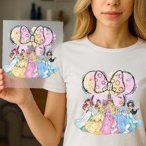 Op de afbeelding: Een wit t-shirt met een grafische print van vijf geanimeerde prinsessen voor een kasteel, bekroond met een grote stippen strikcontour. Het ontwerp maakt gebruik van aquarelachtige spatten in roze, geel en blauw over de illustratie.