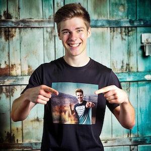 May include: A black crew neck t-shirt featuring a photographic print of a person pointing. The person in the image is also wearing a t-shirt with a graphic. The background is a rustic, teal-colored wooden door.