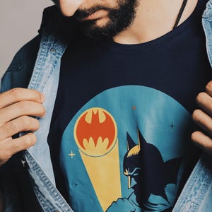 May include: Navy blue t-shirt featuring a Batman graphic with the iconic bat symbol in orange and a silhouette of Batman. The shirt is worn under a denim jacket.