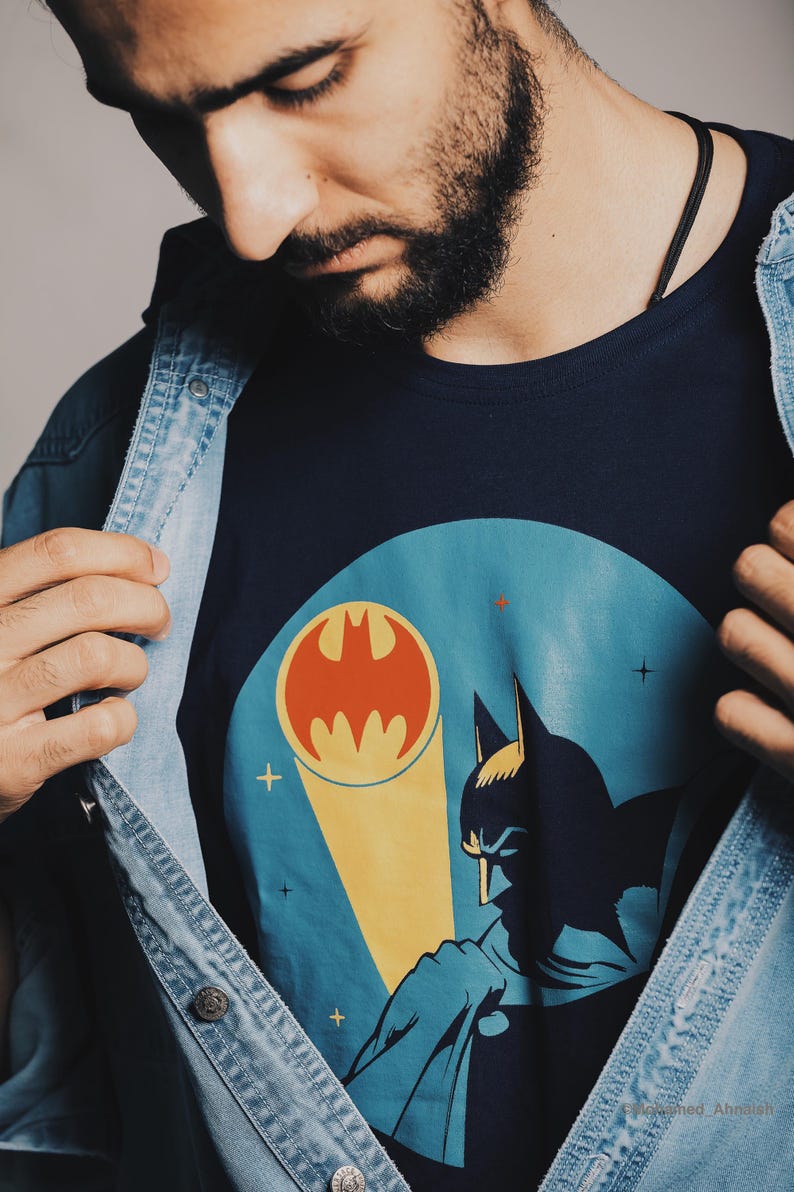 May include: Navy blue t-shirt featuring a Batman graphic with the iconic bat symbol in orange and a silhouette of Batman. The shirt is worn under a denim jacket.