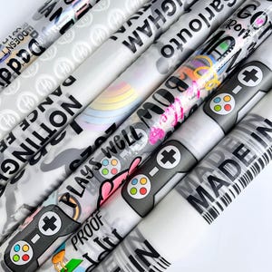 May include: A collection of rolled gift wrap featuring various designs and text. The rolls display text like "NOTTINGHAM", "MADE IN", and "PLAYS WELL". Some rolls have game controller graphics, a rainbow, and a golf scene.