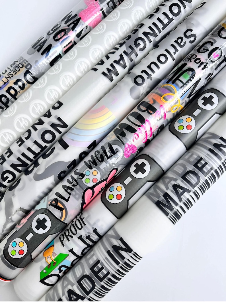 May include: A collection of rolled gift wrap featuring various designs and text. The rolls display text like "NOTTINGHAM", "MADE IN", and "PLAYS WELL". Some rolls have game controller graphics, a rainbow, and a golf scene.