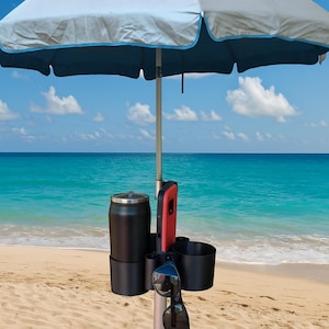 May include: A beach umbrella with a light blue and navy blue canopy, set up on a sandy beach with a turquoise ocean in the background. Attached to the pole is a black drink holder, a red phone holder, and a pair of sunglasses.