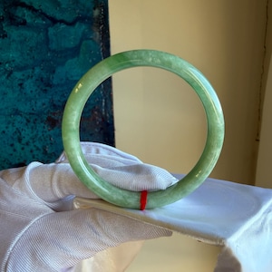May include: A translucent green jade bangle bracelet. The bracelet is a solid, circular band with a smooth, polished surface. A small red string is wrapped around the bracelet. The background is a mix of white fabric and a teal-colored wall.