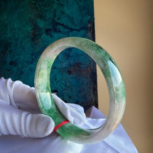 May include: A translucent jade bangle with a gradient of green and white hues. The polished stone is smooth and circular, with a vibrant green section and a lighter, almost clear, section. A red string is visible.