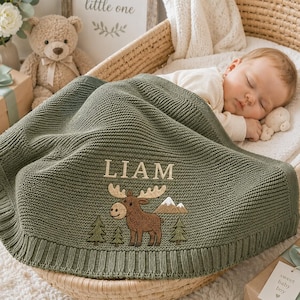 May include: A sage green knitted baby blanket featuring the name LIAM embroidered in white above a brown moose graphic, with small green pine trees and a mountain peak design. The blanket has a ribbed border and is draped over a sleeping infant.