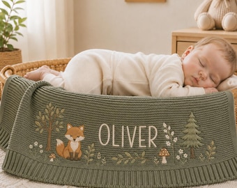May include: A sage green knitted baby blanket featuring an embroidered woodland scene with a fox, trees, mushrooms, and the name Oliver in white lettering. The blanket has a ribbed border and is draped over a wicker basket containing a sleeping baby.