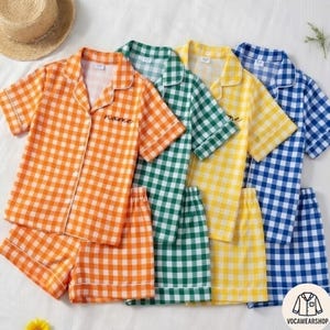 May include: Four sets of short-sleeved pajama sets in orange, green, yellow, and blue gingham patterns. Each set includes a button-down shirt and matching shorts. The word "name" is embroidered on the orange and green shirts. A straw hat is in the background.