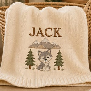 Könnte beinhalten: Eine cremefarbene Strickdecke mit einer Stickerei, die einen grauen Wolfswelpen, zwei grüne Tannen und ein Gebirge zeigt. Der Name JACK ist in braunem Garn über dem Bergmotiv eingestickt. Die Decke hat einen gerippten Rand.