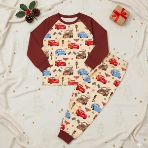 Peut inclure: Un ensemble de pyjama pour enfant avec un fond beige orn&eacute; de motifs de voitures de dessins anim&eacute;s, de feux de circulation et d'&eacute;toiles. Le haut a des manches marron et un col rond. Le pantalon assorti pr&eacute;sente le m&ecirc;me motif de voitures. Un petit coffret cadeau avec un ruban rouge est en arri&egrave;re-plan.