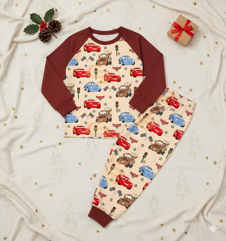 Peut inclure: Un ensemble de pyjama pour enfant avec un fond beige orn&eacute; de motifs de voitures de dessins anim&eacute;s, de feux de circulation et d'&eacute;toiles. Le haut a des manches marron et un col rond. Le pantalon assorti pr&eacute;sente le m&ecirc;me motif de voitures. Un petit coffret cadeau avec un ruban rouge est en arri&egrave;re-plan.