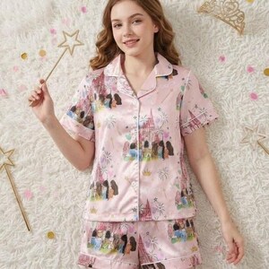 May include: Pink satin pajama set featuring a castle and character print. The short-sleeved top and shorts have white trim. The image also includes a gold crown and star wand, suggesting a princess theme.