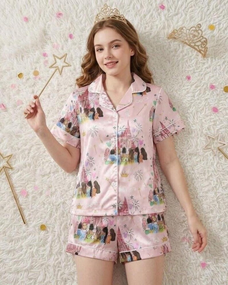 May include: Pink satin pajama set featuring a castle and character print. The short-sleeved top and shorts have white trim. The image also includes a gold crown and star wand, suggesting a princess theme.