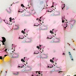 Op de afbeelding: Roze pyjamaset met een herhaald patroon van Minnie Mouse op regenbogen. De top met korte mouwen heeft een kraag en een knoopsluiting. De bijpassende broek maakt de slaapset compleet. Een Minnie Mouse-pop en kleurpotloden zijn ook zichtbaar.
