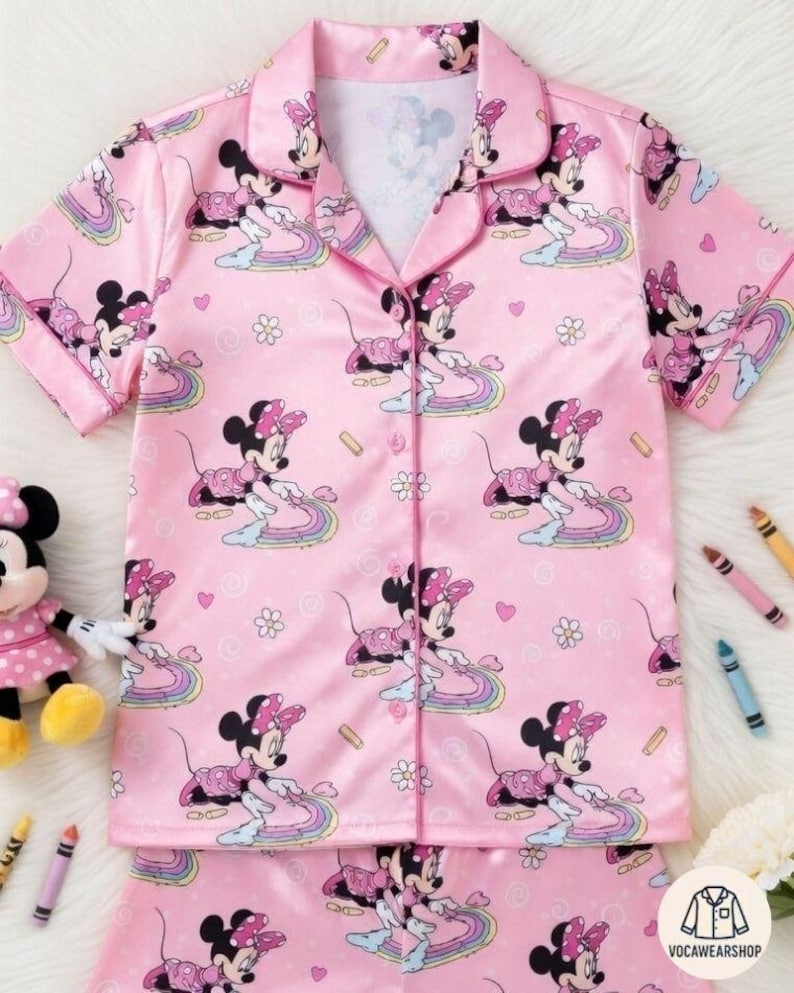Op de afbeelding: Roze pyjamaset met een herhaald patroon van Minnie Mouse op regenbogen. De top met korte mouwen heeft een kraag en een knoopsluiting. De bijpassende broek maakt de slaapset compleet. Een Minnie Mouse-pop en kleurpotloden zijn ook zichtbaar.