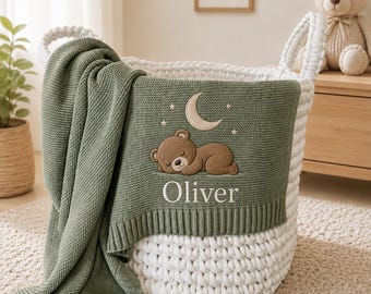 May include: A white chunky knit storage basket containing a sage green knitted blanket. The blanket features an embroidered brown sleeping bear, a crescent moon, small stars, and the name Oliver stitched in white thread on the front.