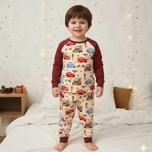 Peut inclure: Un enfant porte un pyjama beige orn&eacute; de voitures de dessins anim&eacute;s rouges, bleues et marron. Le haut &agrave; manches longues a des manches bordeaux et le pantalon des poignets bordeaux. L'enfant sourit et se tient sur un lit.