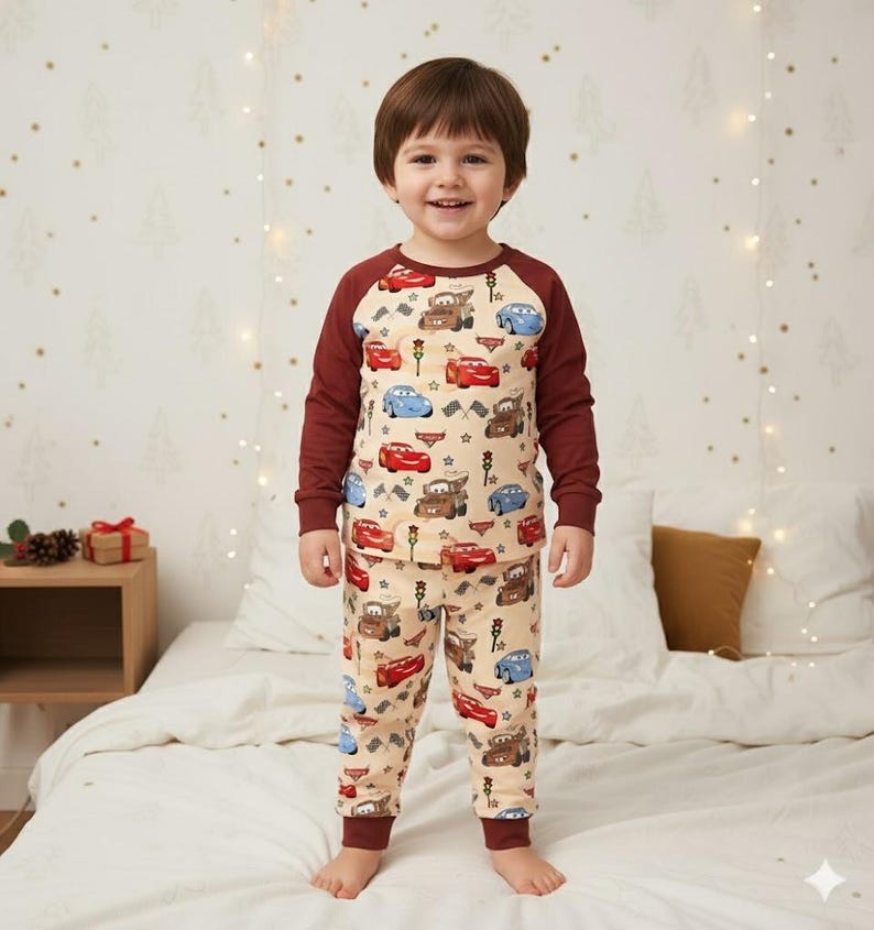 Peut inclure: Un enfant porte un pyjama beige orn&eacute; de voitures de dessins anim&eacute;s rouges, bleues et marron. Le haut &agrave; manches longues a des manches bordeaux et le pantalon des poignets bordeaux. L'enfant sourit et se tient sur un lit.
