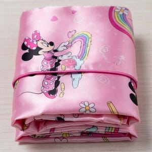 Op de afbeelding: Gevouwen roze satijnen stof met een Minnie Mouse-print, regenbogen, harten en bloemen. De stof is vastgezet met een roze elastiek. Het ontwerp bevat Minnie Mouse in een roze jurk en een stippenstrik.