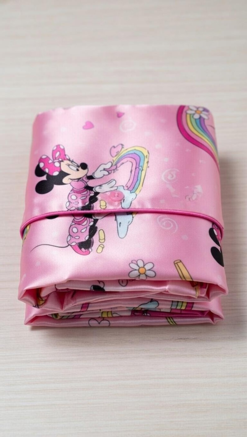 Op de afbeelding: Gevouwen roze satijnen stof met een Minnie Mouse-print, regenbogen, harten en bloemen. De stof is vastgezet met een roze elastiek. Het ontwerp bevat Minnie Mouse in een roze jurk en een stippenstrik.