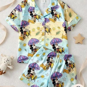 May include: A two-piece pajama set featuring a colorful print of Mickey Mouse relaxing on a beach under an umbrella. The set includes a button-down shirt with short sleeves and matching shorts. The pajamas are in shades of blue, yellow, and purple.