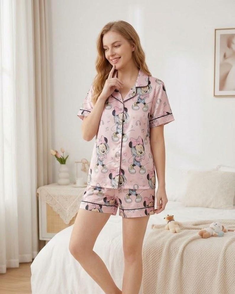 Puede incluir: Conjunto de pijama rosa con estampado de Minnie Mouse. El conjunto incluye una camisa de manga corta con botones y pantalones cortos a juego, ambos con ribete negro. La tela tiene un brillo satinado.