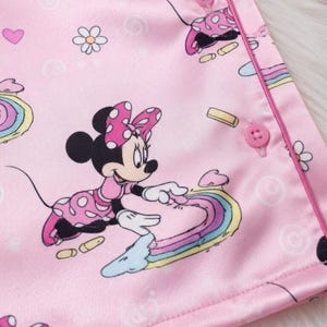 Op de afbeelding: Roze satijnen pyjamastof met Minnie Mouse-illustraties. Het ontwerp bevat Minnie Mouse-figuren, regenbogen, harten en bloemen. De stof heeft een gladde textuur en is afgewerkt met een roze rand en knopen.