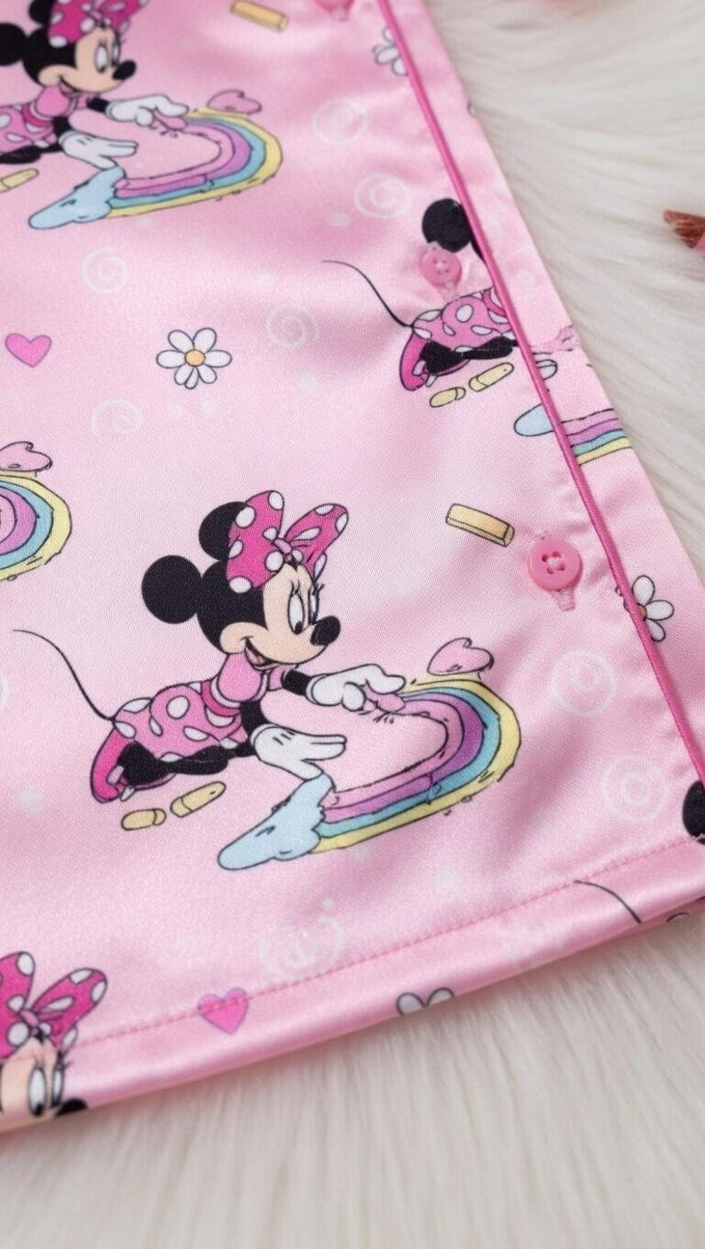 Op de afbeelding: Roze satijnen pyjamastof met Minnie Mouse-illustraties. Het ontwerp bevat Minnie Mouse-figuren, regenbogen, harten en bloemen. De stof heeft een gladde textuur en is afgewerkt met een roze rand en knopen.