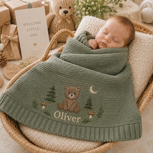 May include: A sage green knitted baby blanket featuring an embroidered brown bear, pine trees, mushrooms, and a crescent moon. The name Oliver is embroidered in white thread at the bottom. The blanket has a ribbed texture and a wide ribbed border.