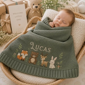 May include: A sage green knitted baby blanket featuring the embroidered name LUCAS in white script, accompanied by detailed illustrations of a fox, a bear, a deer, and a rabbit, all surrounded by small green leaves and tiny flowers.