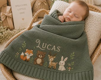 May include: A sage green knitted baby blanket featuring the embroidered name LUCAS in white script, accompanied by detailed illustrations of a fox, a bear, a deer, and a rabbit, all surrounded by small green leaves and tiny flowers.