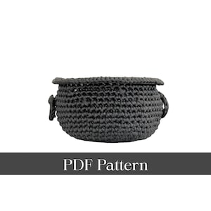 Puede incluir: Una cesta de ganchillo gris oscuro con forma redondeada y dos asas pequeñas. La cesta tiene un aspecto grueso y texturizado y un borde enrollado. El texto "PDF Pattern" se muestra en una pancarta gris oscuro debajo.