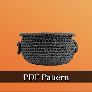 May include: A dark gray, crocheted basket with two small handles. The basket is round with a slightly flared rim. The text "PDF Pattern" is displayed on a brown banner at the bottom of the image. The background is a gradient of orange.