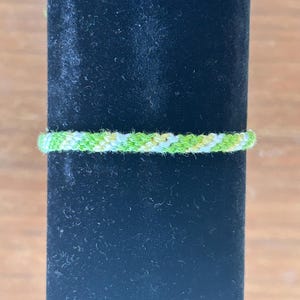 Puede incluir: Una pulsera de la amistad hecha a mano en tonos verdes y blancos. La pulsera tejida se muestra sobre un fondo negro. La pulsera mide aproximadamente 0,6 cm de ancho.