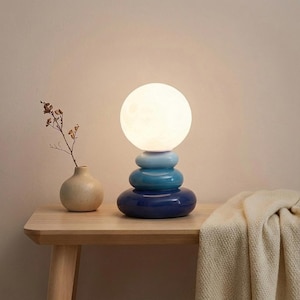 May include: A contemporary table lamp featuring a stacked, rounded base in shades of blue, and a spherical, glowing light. A small beige vase with a sprig of dried flowers sits on a wooden table.