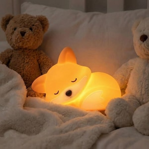 Op de afbeelding: Een gloeiend oranje nachtlampje in de vorm van een vos ligt op een witte deken, geflankeerd door twee teddyberen. Het licht straalt een zachte, warme gloed uit, met zwarte ogen en een wit gezicht. Het nachtlampje is ontworpen om op een slapende vos te lijken.