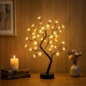 Könnte beinhalten: Eine dekorative baumförmige Lampe mit warmweißen LED-Lichtern in Blütenform. Der schwarze Metallbaum steht auf einem runden Sockel. Eine brennende Kerze und Bücher befinden sich im Vordergrund.