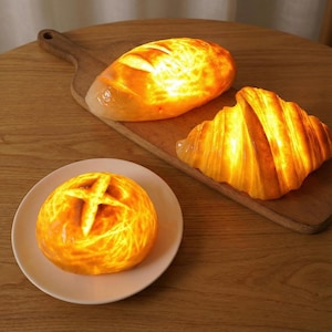 May include: Three illuminated bread-shaped decorative lights. A baguette and a croissant are on a wooden cutting board, and a round loaf is on a white plate. The lights glow with a warm, inviting orange hue.