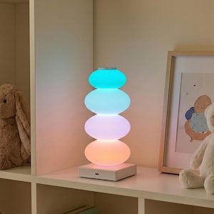 May include: A modern, color-changing LED lamp with a stacked stone design. The lamp emits a soft glow in shades of blue, white, pink, and orange. It sits on a white square base and is ideal for a bedroom or nursery.