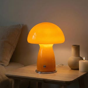 May include: An orange mushroom-shaped table lamp. The lamp has a rounded top and a cylindrical base, with a silver switch. The lamp is on a wooden table next to a beige vase and a lit candle.