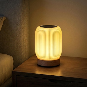 May include: A glowing, cylindrical table lamp with a ribbed, off-white shade. The lamp sits on a round, wooden base and has a dark top. The lamp is on a wooden nightstand next to a bed.