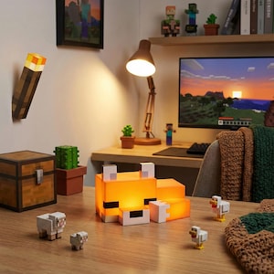 May include: A Minecraft-themed desk setup with a glowing fox lamp, a torch, and character figurines. The lamp gives off a warm orange light. Other items include a wooden chest, cactus, and a computer monitor displaying a Minecraft landscape.