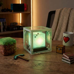 May include: A Minecraft-themed lamp with a glowing green cube inside a clear, pixelated frame. A plush cube, a pickaxe, a mug with a pixelated heart, a treasure chest, and books are also on the wooden surface.