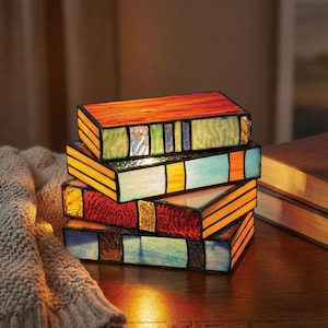 May include: Four stained glass book-shaped boxes in a stack. The boxes feature orange, green, blue, and red panels. They are on a wooden surface. A knitted blanket is in the background. The boxes are reminiscent of a library.