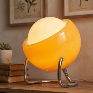 May include: A modern, spherical table lamp with a bright yellow glass shade and a white, glowing interior. The lamp is supported by a curved, chrome-colored metal base. The design is reminiscent of mid-century modern decor.